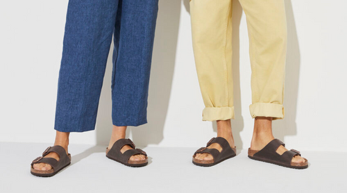 Two people modeling Birkenstock Arizona sandals - one in brown leather paired with blue jeans, the other in black paired with beige pants